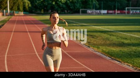 Athlétique jeune femme dans les vêtements de sport courir sur la piste de stade en plein air au coucher du soleil. Concept de condition physique, de détermination et de mode de vie actif. Banque D'Images