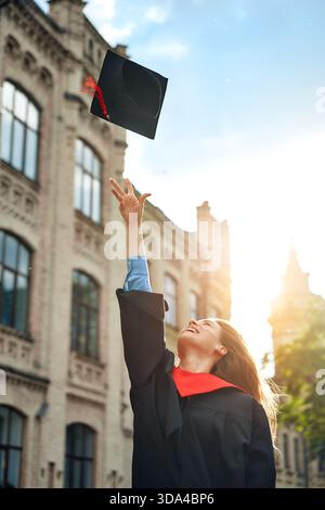 Une diplômée célèbre joyeusement sa réussite scolaire en jetant sa casquette en l'air sur un campus universitaire prestigieux, symbolisant le succès A. Banque D'Images