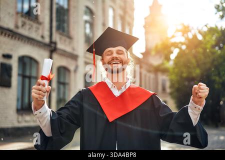 Un jeune homme en casquette et robe célèbre son succès académique en plein air, tenant fièrement son diplôme à la lumière du soleil, exprimant fierté et bonheur pour Banque D'Images
