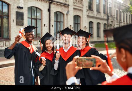 Divers diplômés en casquettes et robes célèbrent joyeusement à l'extérieur d'un bâtiment historique, posant pour des photos après leur cérémonie de remise des diplômes, symbolisant acad Banque D'Images