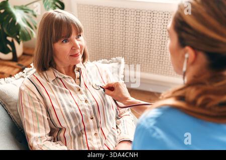 Femme médecin de famille à la maison en uniforme médical avec stéthoscope vérifiant la fréquence cardiaque de la femme plus âgée assise sur le canapé dans le salon. Les soins de santé et moi Banque D'Images