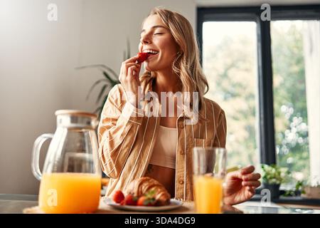 Jeune femme heureuse assise à table dans la cuisine, mangeant des croissants avec des fraises et buvant du jus. Femme prenant le petit déjeuner dans un kitche confortable Banque D'Images