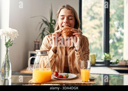 Jeune femme heureuse assise à table dans la cuisine, mangeant des croissants avec des fraises et buvant du jus. Femme prenant le petit déjeuner dans un kitche confortable Banque D'Images