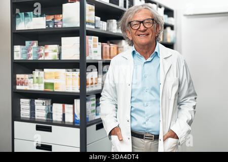 Pharmacien senior souriant en manteau blanc et lunettes debout dans la pharmacie près des étagères avec des médicaments, à l'air confiant et professionnel Banque D'Images