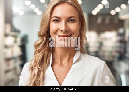Portrait d'une pharmacienne confiante avec de longs cheveux blonds souriant à la caméra tout en se tenant dans une pharmacie moderne. Concept : soins de santé, personnalisé Banque D'Images