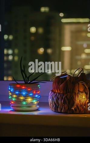 Plante en pot enveloppée de guirlandes lumineuses colorées sur le rebord de la fenêtre la nuit, avec des lumières de ville floues en arrière-plan. Décoration de maison confortable pour les vacances Banque D'Images