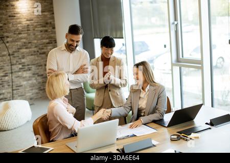 Deux professionnels se serrent la main dans un bureau lumineux. Un groupe à proximité montre de l'enthousiasme et du soutien pour l'accord réussi. Cette scène capture Teamwo Banque D'Images