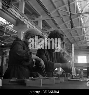 Slavyansk, RSS d'Ukraine, URSS - années 1970 : deux travailleuses en foulards et vêtements de travail dans l'atelier d'assemblage de l'usine de Stroymash mesurant des pièces métalliques. Les femmes utilisent une jauge de hauteur et des étriers pour vérifier la qualité des composants mécaniques sur un établi. Cette photo d'archives en noir et blanc capture l'atmosphère de la main-d'œuvre féminine qualifiée et la puissance industrielle de la paisible région du Donbass à l'époque soviétique. Employés du service de contrôle qualité au travail. Banque D'Images
