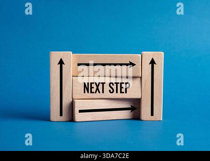 Blocs en bois affichant le texte de L'ÉTAPE SUIVANTE avec des flèches, symbolisant le choix de la direction, de la progression, de la stratégie et du développement en avant sur un fond bleu Banque D'Images