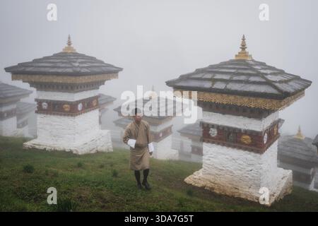 Vue d'un homme en tenue traditionnelle bhoutanaise marche parmi les stupas Dochula Chorten, enveloppés de brume, leurs toits dorés brillants doucement, Hungtso, B. Banque D'Images