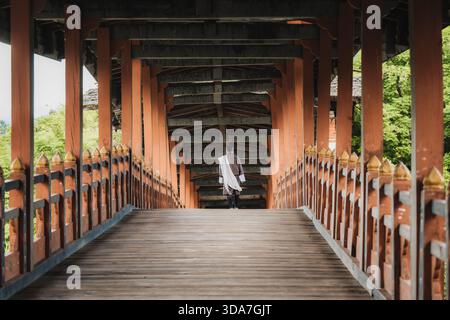 Vue d'une figure solitaire drapée de tissu blanc coulant marche le long d'un pont en bois avec des balustrades orange vives, encadré par une structure en bois symétrique Banque D'Images