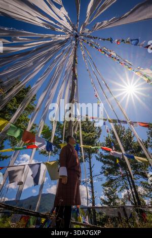 Vue de drapeaux de prière flottant autour d'un haut poteau sous un soleil éclatant, avec une personne en tenue traditionnelle observant d'en bas, col de Kikila, Bumthan Banque D'Images