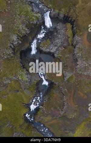 Vue aérienne d'une cascade en cascade sculptant à travers le terrain accidenté et couvert de mousse, ses eaux vives contrastant fortement avec le sombre volcanique R Banque D'Images