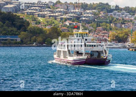Istanbul, Turquie - 24 août 2025 : un ferry de passagers blanc et violet glisse sur l'eau bleue vibrante de Bodphorus tandis qu'un quartier à flanc de colline avec t Banque D'Images