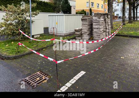 Du ruban de sécurité rouge et blanc délimite une zone de construction dans une rue. Les matériaux en carton sont empilés près d'une boîte utilitaire. La scène montre de l'herbe verte et une surface de béton sous un ciel nuageux. Banque D'Images