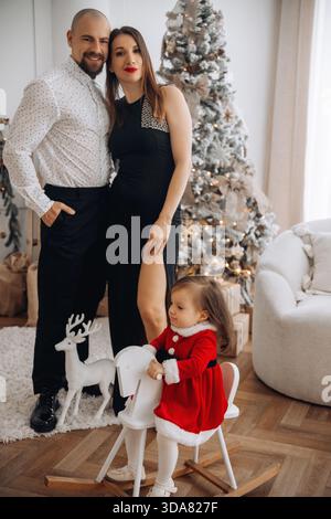 La famille se tient ensemble dans un salon avec un arbre de Noël. L'enfant en tenue rouge est assis sur une petite chaise. Les parents portent des vêtements formels et smil Banque D'Images