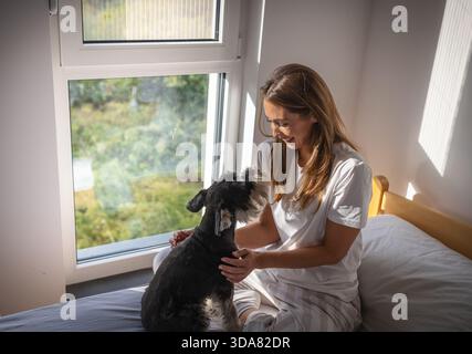 Une femme interagit avec un chien assis sur un lit près d'une grande fenêtre. La lumière du soleil remplit la pièce, créant une atmosphère matinale lumineuse Banque D'Images