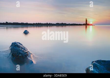 Scène de crépuscule sereine avec un littoral calme avec des fonds marins visibles, menant à un brise-lames lointain et un phare silhouette sur un doux coucher de soleil Banque D'Images