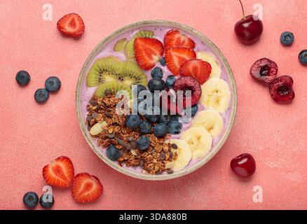 Bol d'açai vibrant et nutritif surmonté de tranches de kiwi, fraises, bleuets, cerises, bananes, et le granola, un repas coloré et sain ou un petit déjeuner sur un fond rose Banque D'Images