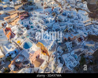 Vue aérienne des églises emblématiques au dôme bleu nichées parmi les bâtiments blanchis à la chaux accrochés aux falaises, scintillant sous la lumière dorée, Oia, Banque D'Images