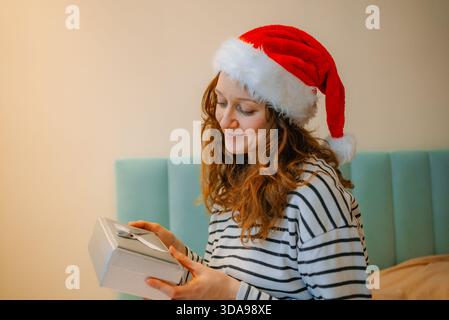 Femme en chapeau de père noël et pyjama ouvrant un cadeau sur son lit, expérimentant la joie de recevoir un cadeau de noël avec une expression heureuse et ravie Banque D'Images
