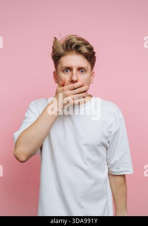 Jeune homme surpris couvrant la bouche avec la main sur fond rose. Banque D'Images