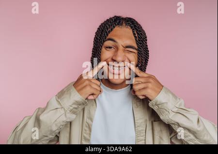 Adolescent souriant avec des accolades, pointant vers les joues, fond rose. Banque D'Images