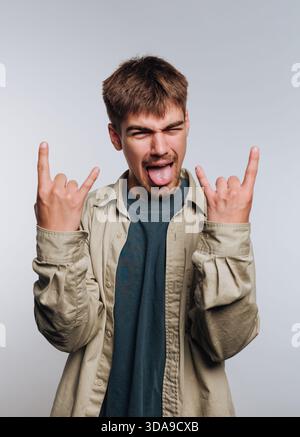 Un jeune homme sourit et sort sa langue tout en faisant un symbole de roche avec ses mains. Il porte une chemise légère sur un t-shirt sombre et est à l'intérieur agai Banque D'Images