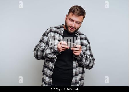 Un jeune homme dans une chemise à carreaux se tient à l'intérieur, concentré sur son smartphone comme il aime jouer. Il semble détendu et engagé dans son activité, smilin Banque D'Images