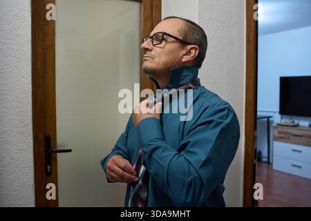Un homme mûr portant une chemise à col bleu foncé et des lunettes à bordure noire ajuste soigneusement sa cravate à motifs Banque D'Images