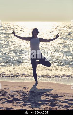 Jeune femme adulte effectue le yoga de pose d'arbre sur la plage de sable en scintillant la mer Baltique. La scène paisible rétroéclairée évoque la pleine conscience et l'équilibre au soleil chaud Banque D'Images