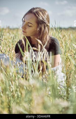 Jeune femme avec les cheveux bruns et chemise noire est assis paisiblement parmi de grandes tiges de seigle sous le ciel clair. Un éclairage naturel lumineux améliore la sérénité de l'envir rural Banque D'Images