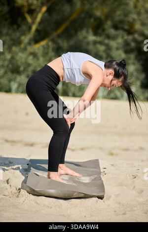 Femme adulte en leggings noirs et débardeur blanc se plie en avant dans la pose de yoga sur tapis gris par Sandy Beach. Fond naturel calme avec feuillage vert com Banque D'Images
