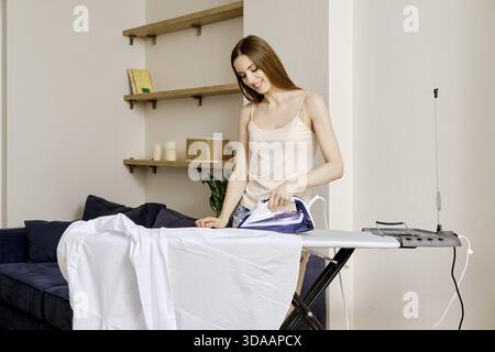 Une femme se tient à côté d'une planche à repasser dans un salon lumineux. Elle presse une chemise blanche avec un fer à repasser. La chambre a des étagères avec des livres et décoratio Banque D'Images
