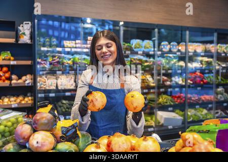 Femme employée d'épicerie portant un tablier et des gants, souriant tout en tenant deux grenades, arrangeant des fruits sains pour la consommation fraîche sur un Banque D'Images