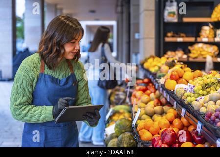 Femme dans un tablier et des gants de gestion des stocks, vérifier les niveaux de stock de fruits frais et de produits sur une tablette numérique dans une petite épicerie stor Banque D'Images