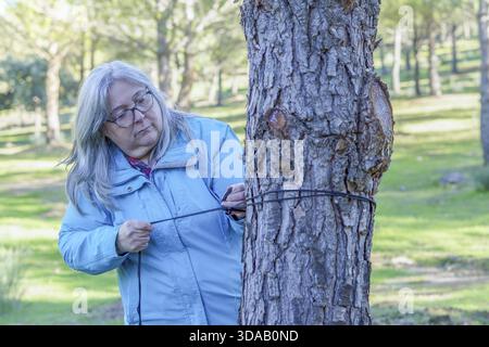 Femme senior portant des lunettes et une veste bleue attachant une corde noire autour d'un tronc d'arbre dans un parc public Banque D'Images