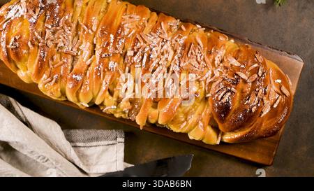Une photo en gros plan, à angle élevé, de cuisine sombre montre un pain sucré tressé fraîchement cuit, brillant, arrosé de glaçage et recouvert d'amandes tranchées Banque D'Images