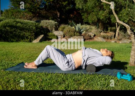 Femme âgée allongée sur un tapis d'exercice tout en massant les muscles du dos avec des rouleaux en mousse au parc, en profitant de l'entraînement thérapeutique et rafraîchissant, et le bien-être dans un cadre naturel Banque D'Images