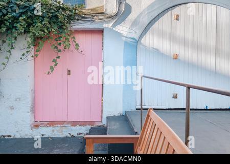 Détail d'une porte en bois rose à côté d'une grande porte de garage en bois voûté blanc, avec lierre grimpant et une chaise en bois au premier plan Banque D'Images