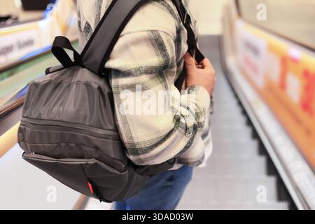Gros plan d'un homme avec un sac à dos gris debout sur l'escalier roulant. Personne portant une chemise à carreaux dans un centre commercial ou à l'aéroport. Photo de haute qualité Banque D'Images