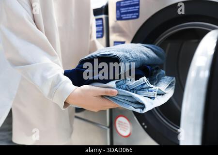 Femme tenant pile de jeans propres près de la machine à laver. Gros plan des mains avec la lessive à la laverie automatique. Photo de haute qualité Banque D'Images