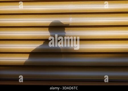 Ombre artistique d'un homme portant une casquette coulée sur un store à rayures jaunes. L’image joue avec la lumière, la géométrie et le minimalisme Banque D'Images