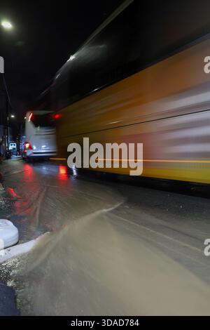 Photo longue exposition d'un bus jaune et blanc conduisant rapidement dans une rue de la ville baignée de pluie la nuit. Banque D'Images