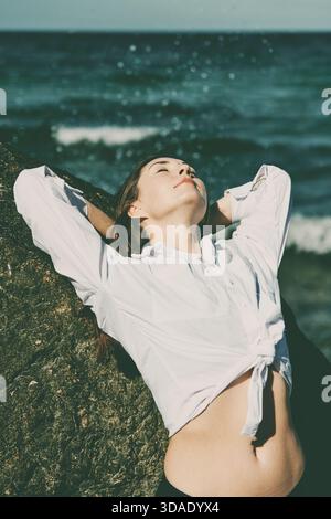 Jeune femme en chemise blanche se penche contre le rocher, les yeux fermés, le visage tourné vers le haut vers le soleil. Les vagues de la mer Baltique et l'eau bleu-vert forment le bac naturel Banque D'Images