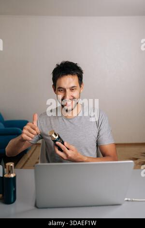 Vlogger masculin joyeux donnant les pouces vers le haut et tenant le produit cosmétique pendant un appel vidéo en ligne, examinant les articles de beauté pour le public des médias sociaux ou en direct Banque D'Images