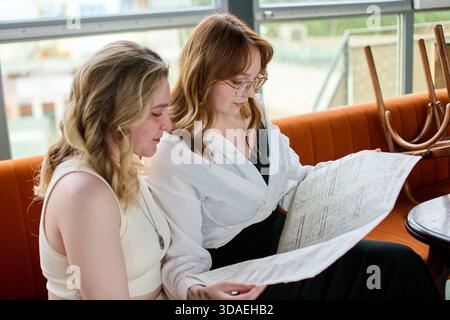 Deux jeunes femmes avec la peau claire et les cheveux, une blonde et une rousse avec des lunettes, assis sur le canapé orange dans le café, concentré sur la lecture grand menu sous na Banque D'Images