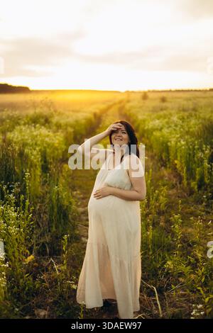 Une femme enceinte souriante marche à travers un champ ensoleillé, touchant doucement son ventre. Son expression reflète le bonheur et l’anticipation, entourée de lu Banque D'Images