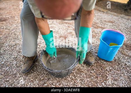 Image recadrée méconnaissable d'un homme senior anonyme mélangeant du ciment dans un seau noir à l'aide d'une truelle. Il porte des gants verts, se concentrant sur une sortie Banque D'Images