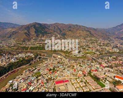 Vue aérienne de la ville tentaculaire nichée entre la rivière sinueuse et les montagnes imposantes sous un ciel clair, Muzaffarabad, Cachemire, Pakistan. Banque D'Images